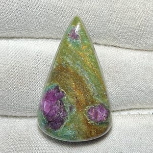 May include: A polished, teardrop-shaped gemstone with a green and gold base colour. The stone features vibrant, irregular patches of deep pink and purple hues. The gem is smooth and appears to be set against a neutral background.
