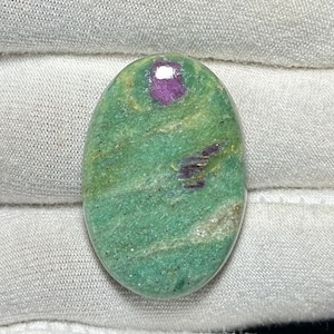 Puede incluir: Gema ovalada con una base verde vibrante, salpicada de tonos verdes y beige más oscuros. La piedra presenta una prominente inclusión magenta oscura en la parte superior y pequeñas inclusiones magenta.