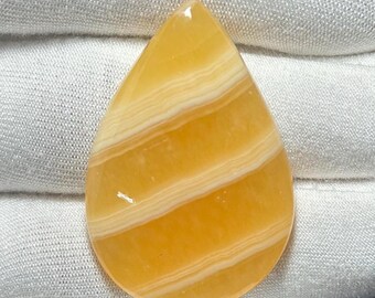 May include: A teardrop-shaped gemstone with a polished surface. The stone is a warm yellow color with horizontal bands of creamy white. The gemstone is set against a white fabric background.
