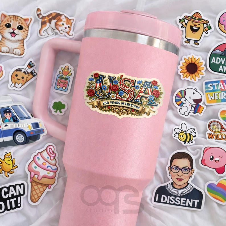 Puede incluir: Un vaso t&eacute;rmico rosa adornado con varias pegatinas. Las pegatinas incluyen un dise&ntilde;o de EE. UU. con el texto "250 YEARS OF FREEDOM", personajes de dibujos animados y otros gr&aacute;ficos. El vaso tiene un borde plateado y un asa grande.