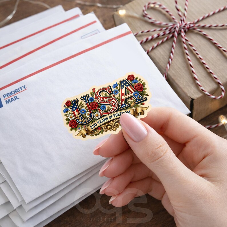 Puede incluir: Una pegatina con la palabra "USA" en rojo, blanco y azul, decorada con flores y el texto "250 Years of Freedom". La pegatina est&aacute; sujeta en una mano, con sobres blancos y una caja de regalo en el fondo.