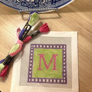 K&ouml;nnte beinhalten: Eine Gobelin-Leinwand mit einem gr&uuml;nen Zentrum und einem rosa Buchstaben "M", umgeben von einem lila Rahmen. Stickgarne in Pink, Gr&uuml;n und Lila sind zusammengebunden. Eine dekorative Platte befindet sich im Hintergrund.