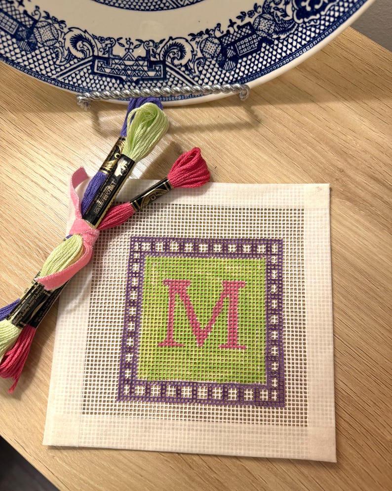 K&ouml;nnte beinhalten: Eine Gobelin-Leinwand mit einem gr&uuml;nen Zentrum und einem rosa Buchstaben "M", umgeben von einem lila Rahmen. Stickgarne in Pink, Gr&uuml;n und Lila sind zusammengebunden. Eine dekorative Platte befindet sich im Hintergrund.