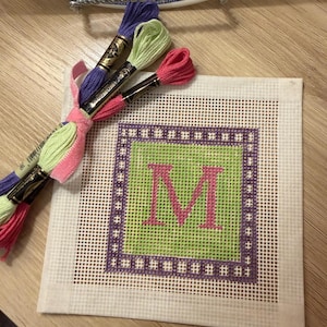 K&ouml;nnte beinhalten: Ein quadratischer Nadelkissenstoff mit einem gr&uuml;nen Zentrum und dem rosa Buchstaben "M". Der Stoff ist von einem lila Rahmen umgeben. Geb&uuml;ndeltes Stickgarn in Rosa, Gr&uuml;n und Lila ist mit einem rosa Band gebunden.