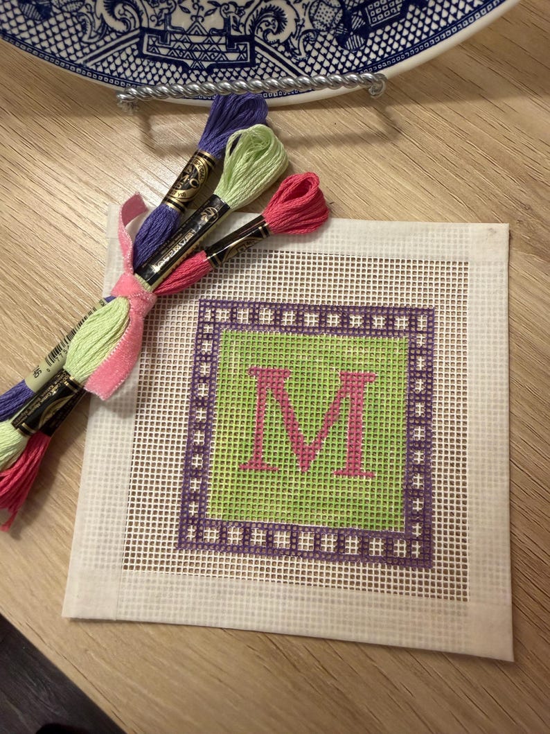K&ouml;nnte beinhalten: Ein quadratischer Nadelkissenstoff mit einem gr&uuml;nen Zentrum und dem rosa Buchstaben "M". Der Stoff ist von einem lila Rahmen umgeben. Geb&uuml;ndeltes Stickgarn in Rosa, Gr&uuml;n und Lila ist mit einem rosa Band gebunden.