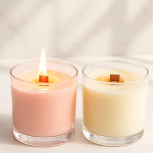 May include: Two glass jar candles with wooden wicks. The candle on the left is light pink and lit with a bright flame, while the candle on the right is cream-colored and unlit. Both candles are contained in clear, cylindrical glass vessels.