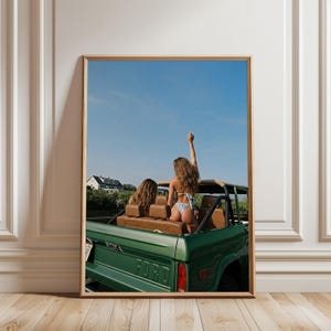 May include: A framed wall art print depicting two individuals in a green vintage Ford Bronco convertible. One individual stands in the back, wearing a light-colored bikini, with one arm raised against a clear blue sky. The vehicle has brown seats.