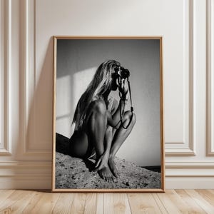 Könnte beinhalten: Ein schwarz-weiß gerahmter Wandkunstdruck, der eine Person mit langem Haar zeigt, die hockt und eine Vintage-Kamera vor das Gesicht hält. Die Komposition nutzt kontrastreiches Licht, um die Figur vor einem schlichten Hintergrund hervorzuheben.