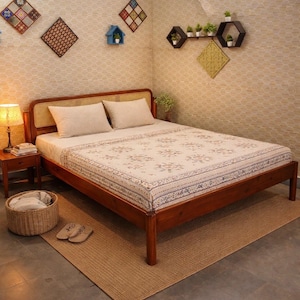 Puede incluir: Una cama de madera con una colcha estampada floral y dos almohadas. La habitación tiene adornos decorativos en la pared y una cesta de mimbre. Una pequeña mesita de noche con una lámpara está al lado de la cama. La cama mide unos 200 cm de largo.