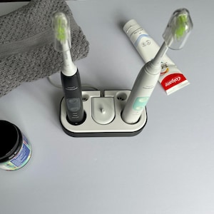 May include: An overhead shot of a dental hygiene setup. It includes two electric toothbrushes in a charging base, a tube of Colgate toothpaste, a jar with a black lid, and a gray washcloth. The toothbrushes have green brush heads.