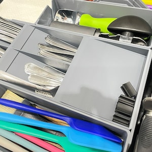 May include: A gray drawer organizer filled with silverware, serving utensils, and kitchen tools. Silver spoons and forks are neatly arranged in separate compartments. Colorful spatulas and a bottle opener are also visible.
