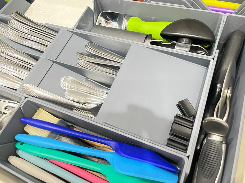 May include: A gray drawer organizer filled with silverware, serving utensils, and kitchen tools. Silver spoons and forks are neatly arranged in separate compartments. Colorful spatulas and a bottle opener are also visible.