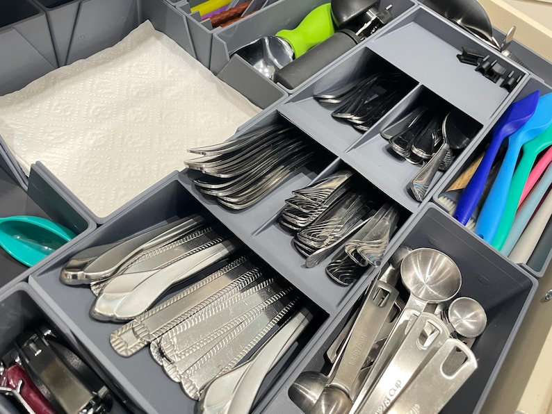 May include: A neatly organized drawer with gray dividers, filled with silverware, serving spoons, and measuring spoons. A stack of white paper towels is visible. Colorful plastic spoons and spatulas add a pop of color to the arrangement.