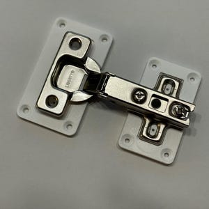 May include: A cabinet hinge with a silver finish and white mounting plates. The hinge is made of metal and has the brand name "LIBERTY" stamped on it. The mounting plates have pre-drilled holes for screws.