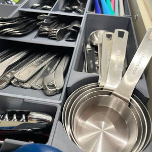 May include: A kitchen drawer organizer filled with silverware, measuring cups, and a corkscrew. The measuring cups are stainless steel and labeled with cup and milliliter measurements. The silverware includes forks, spoons, and knives.