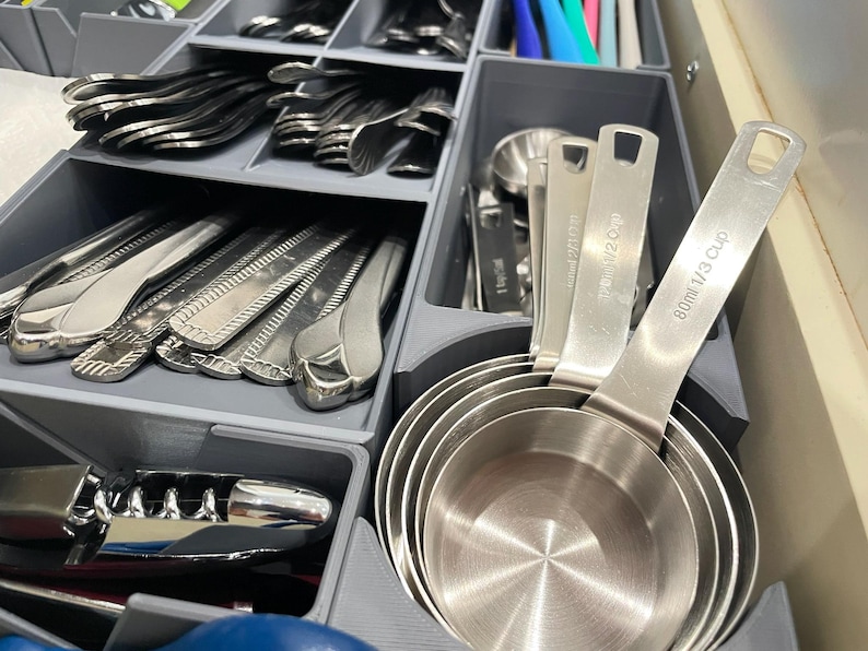 May include: A kitchen drawer organizer filled with silverware, measuring cups, and a corkscrew. The measuring cups are stainless steel and labeled with cup and milliliter measurements. The silverware includes forks, spoons, and knives.