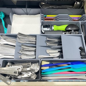 May include: A kitchen drawer organized with gray dividers, filled with silverware, measuring cups, and various kitchen utensils. Includes spoons, forks, knives, spatulas, and a teal scoop. The drawer is neatly arranged for easy access and storage.