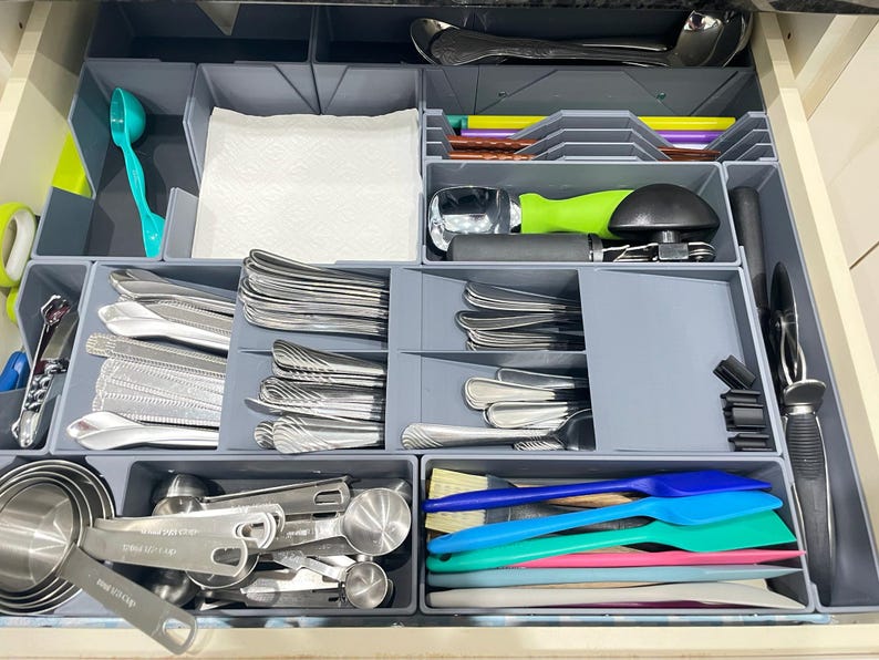 May include: A kitchen drawer organized with gray dividers, filled with silverware, measuring cups, and various kitchen utensils. Includes spoons, forks, knives, spatulas, and a teal scoop. The drawer is neatly arranged for easy access and storage.