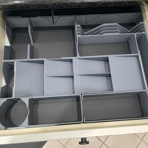 May include: A gray plastic drawer organizer with multiple compartments of varying sizes and shapes. The organizer is designed to fit inside a drawer and features rectangular, square, and circular sections for storing items. The drawer is open, revealing the organizer.