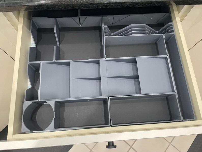 May include: A gray plastic drawer organizer with multiple compartments of varying sizes and shapes. The organizer is designed to fit inside a drawer and features rectangular, square, and circular sections for storing items. The drawer is open, revealing the organizer.