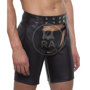 May include: Black leather shorts featuring a high-waisted design with a front snap-button closure and a vertical zipper running down the center. The garment has an open-front crotch area and includes a circular logo with the text RA LEATHERS.