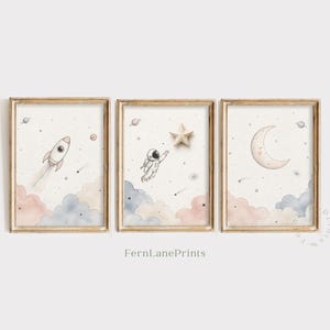 May include: Three framed wall art prints featuring watercolor space illustrations. The left print shows a rocket, the center shows an astronaut reaching for a star, and the right shows a crescent moon. All prints include pastel clouds and stars.