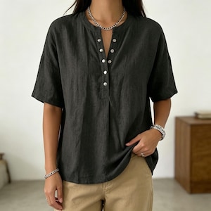 May include: Dark gray short-sleeve shirt with a mandarin collar and a button-down placket. The shirt has a relaxed fit and is made of a textured fabric. The model is wearing the shirt with khaki-colored pants and silver jewelry.