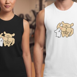 Pode incluir: Camisetas sem mangas pretas e brancas com uma ilustração de desenho animado de um cão musculoso flexionando o bíceps ao lado de um personagem de desenho animado branco menor. A imagem mostra duas pessoas em uma academia.