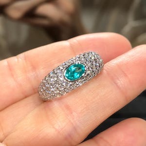 May include: A silver-toned ring featuring a central oval-cut bright teal gemstone surrounded by a pavé setting of numerous small, clear, round-cut stones that cover the entire visible surface of the wide, domed band of the jewellery piece.