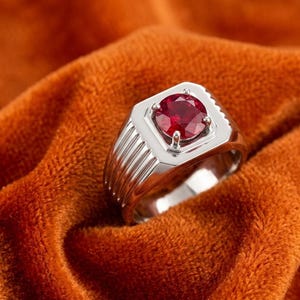 Puede incluir: Un anillo de tono plateado con una piedra preciosa roja profunda, redonda y facetada, engastada en un bisel cuadrado. La banda del anillo tiene un acabado pulido con detalles de ranuras verticales en los lados, creando un aspecto metálico estructurado.