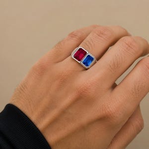 Peut inclure: Une bague argentée ornée de deux pierres précieuses rectangulaires côte à côte. L'une est d'un rouge éclatant et l'autre d'un bleu profond. L'anneau en métal présente une finition lisse et polie, avec un sertissage aux lignes épurées.