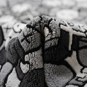May include: A tufted rug featuring a monochromatic gray, black, and white graphic pattern with bold outlines and abstract shapes. The textile has a thick, textured pile surface that is folded over to reveal the dense, looped construction of the fabric.
