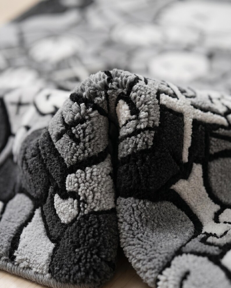 May include: A tufted rug featuring a monochromatic gray, black, and white graphic pattern with bold outlines and abstract shapes. The textile has a thick, textured pile surface that is folded over to reveal the dense, looped construction of the fabric.