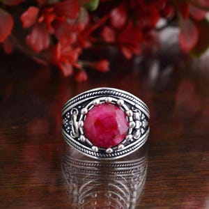 May include: A silver ring with a large, round, deep red gemstone at the center. The ring has intricate detailing around the gemstone and along the band, with a textured pattern and small silver beads. The background features blurred red flowers.