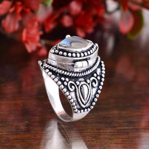 Op de afbeelding: Een zilveren ring met een sierlijk ontwerp. De ring heeft een vierkante, iriserende steen gezet in een gedetailleerde bezel. De band heeft een decoratief patroon van stippen en krullen. De ring wordt gepresenteerd op een houten oppervlak.