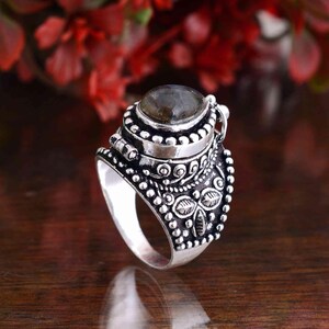 May include: A silver ring with an oval, gray gemstone. The ring features intricate detailing, including small beads and floral patterns. The band is wide and the setting is ornate, creating a vintage or bohemian aesthetic.