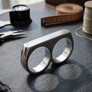 May include: A polished silver two-finger ring with a geometric pattern on the top. The ring features two circular openings for the fingers. It is displayed on a dark surface, with tools and a measuring tape in the background. The ring's design is modern.