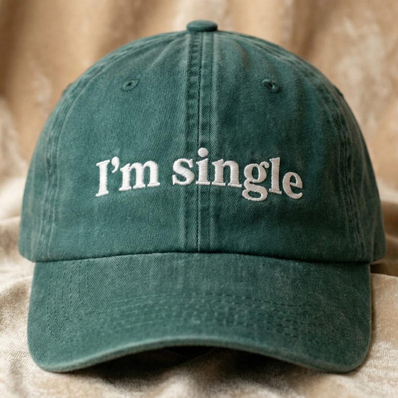 May include: A green baseball cap with the embroidered text "I'm single" in white. The cap has a curved brim and a button on top. The hat is made of a soft, textured fabric.