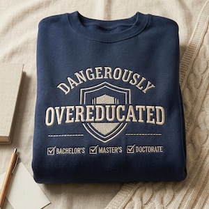 May include: Navy blue sweatshirt with the embroidered text "DANGEROUSLY OVEREDUCATED" in cream-colored thread. The design includes a shield and checkmarks for Bachelor's, Master's, and Doctorate degrees. The sweatshirt is folded.
