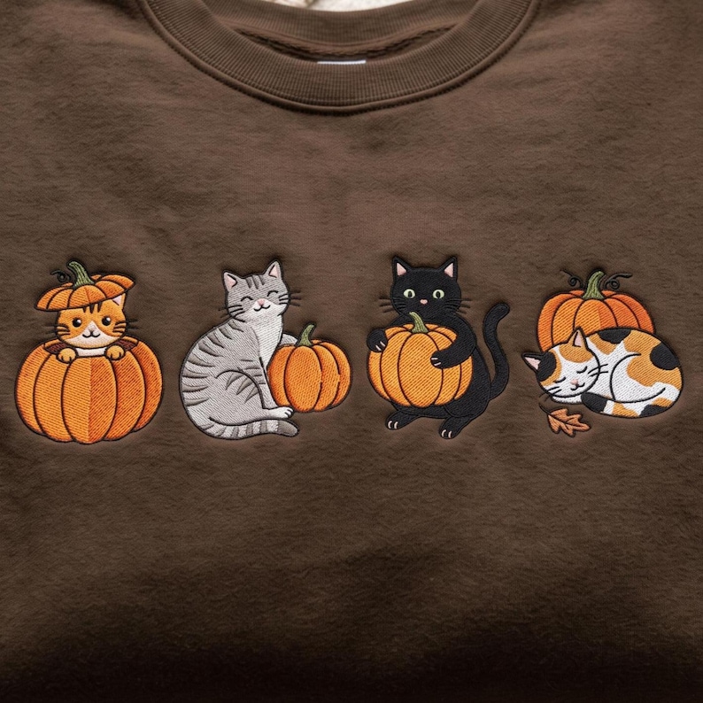 May include: A brown sweatshirt featuring an embroidered design of four cats with pumpkins. The cats include an orange tabby inside a pumpkin, a gray tabby, a black cat holding a pumpkin, and a calico cat sleeping next to a pumpkin.