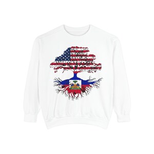Puede incluir: Una sudadera blanca de manga larga con un gráfico de un árbol. La copa del árbol muestra el patrón de la bandera de los Estados Unidos, mientras que las raíces y el tronco presentan el diseño de la bandera de Haití, incluyendo el escudo central.