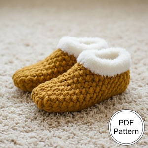 May include: A pair of mustard yellow crochet slippers with a fluffy white trim. The slippers are made of a thick, textured yarn and are sitting on a cream-colored carpet. A white circle with the text "PDF Pattern" is in the lower right corner.