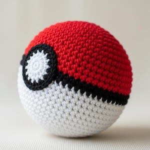 Puede incluir: Una bola de ganchillo en rojo y blanco, con una franja negra y un diseño circular en blanco y negro. La bola hecha a mano es una esfera, con una superficie texturizada. El juguete es un diseño popular.