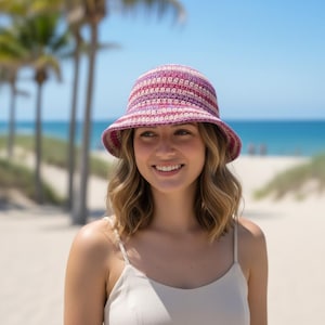 Könnte beinhalten: Häkel-Eimerhut in Rosa- und Lilatönen mit breiter Krempe. Der Hut hat horizontale Streifen in verschiedenen Rosa- und Lilatönen. Der Hintergrund zeigt eine Strandszene mit Palmen und dem Meer.