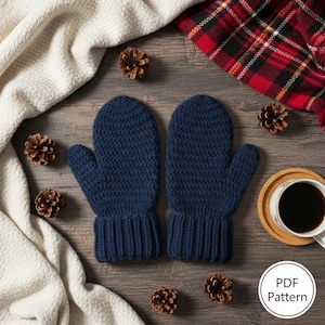 May include: A pair of navy blue knitted mittens with ribbed cuffs, displayed on a wooden surface with pine cones, a white scarf, and a red plaid blanket. A cup of coffee sits on a wooden coaster. The image includes a PDF pattern icon.