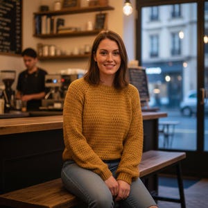 May include: A woman wearing a mustard yellow crochet sweater and blue jeans sits on a wooden bench inside a cafe. The cafe has a warm, inviting atmosphere with soft lighting and a barista working behind the counter.