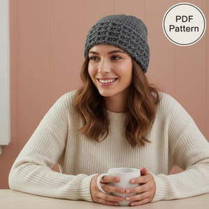 Op de afbeelding: Een grijze gehaakte beanie met een textuurpatroon. De beanie wordt gedragen door een persoon die een crèmekleurige trui draagt. Een witte mok wordt in de handen van de persoon gehouden. De afbeelding bevat een cirkelvormige afbeelding met de tekst "PDF Pattern".