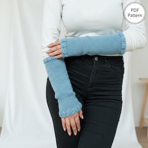 May include: A pair of light blue knitted arm warmers. The arm warmers are a solid color with a ribbed cuff and are worn by a person wearing a white long-sleeve shirt and black jeans. The image includes the text "PDF Pattern".