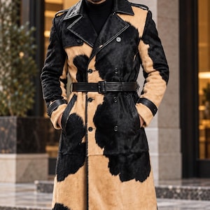 May include: A double-breasted trench coat featuring a cow-print pattern in black and tan fur. The coat includes a black leather belt with a buckle, black leather lapel trim, and black buttons. The text MontreuxLeather is visible in the top corner.