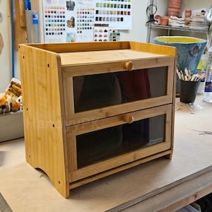 May include: A light brown bamboo storage unit featuring two horizontal drawers with clear glass panels and small round wooden knobs. The structure has a flat top surface and side panels with visible circular cutouts, providing a natural wood finish.